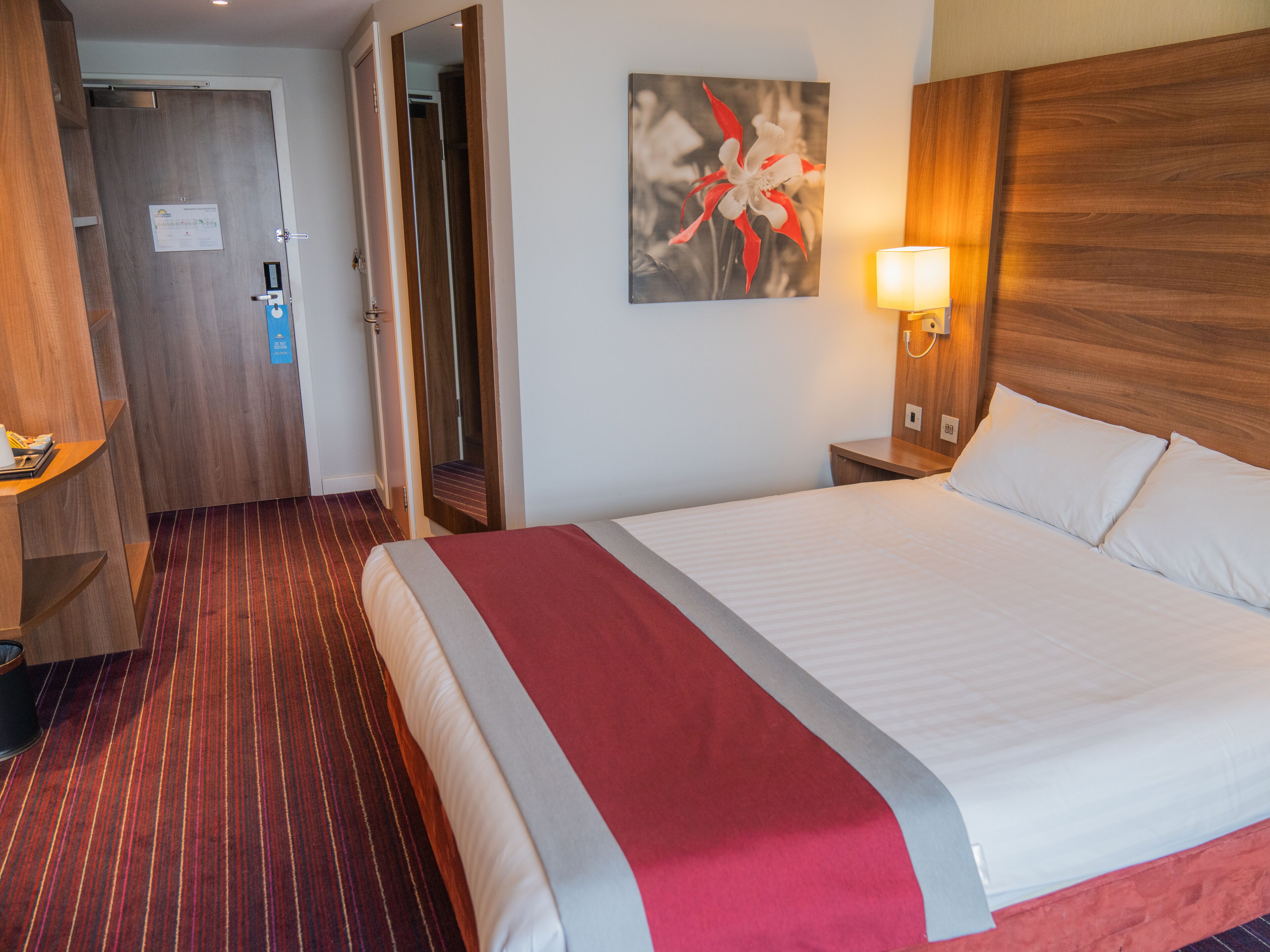 A hotel room with a large bed, a wooden headboard, a bedside table with a lamp, a mirror, and a red-striped carpet.