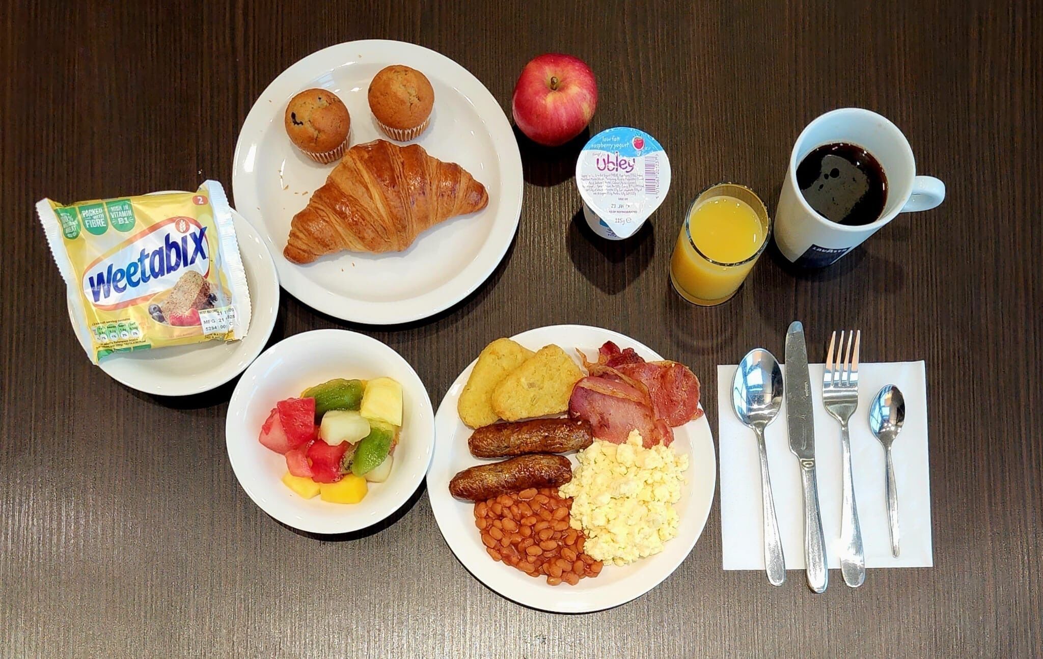 A breakfast table with various food items including Weetabix, croissants, an apple, yogurt, orange juice, coffee, sausages, beans, scrambled eggs, and a bowl of fruit.