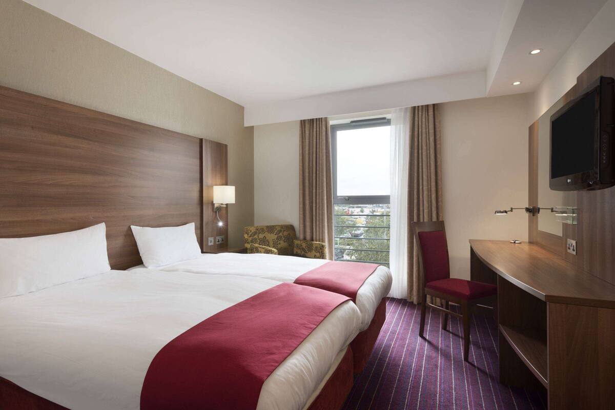 A hotel room with two beds, a desk, a TV, and a view of the outdoors.