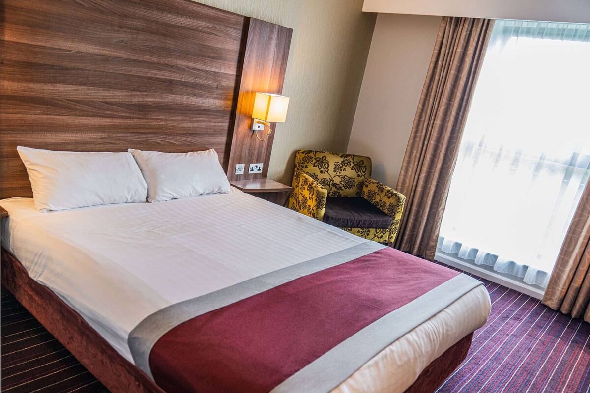 A hotel room with a large bed, a nightstand with a lamp, a floral armchair, and a window with curtains.