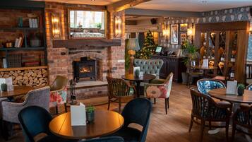 A cozy pub with brick walls, a fireplace, and a Christmas tree. There are tables and chairs for dining, and a bar area with wooden cabinets.