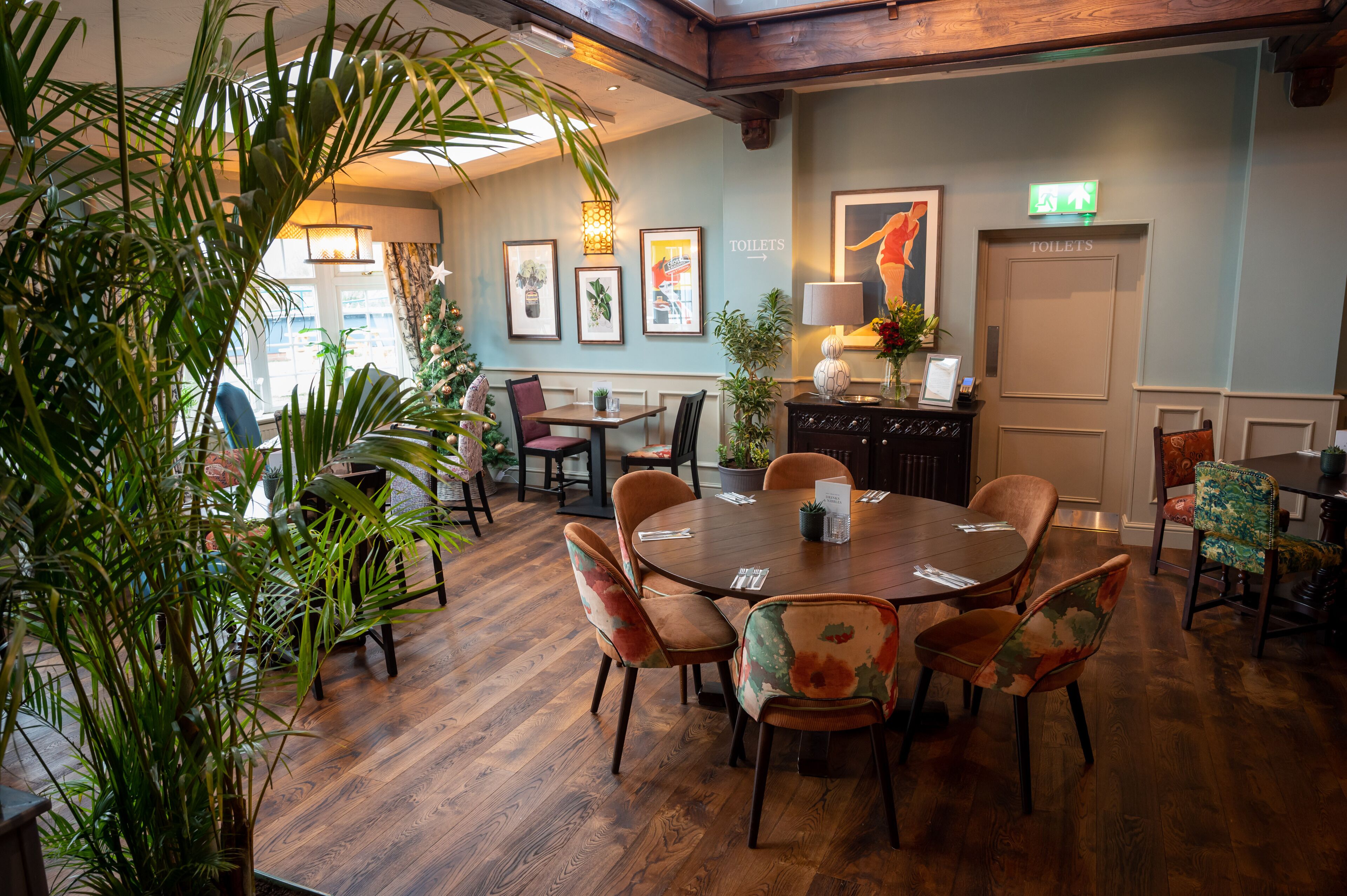 A dining area with wooden flooring, a central round table, and floral-patterned chairs. There are framed pictures on the wall, a potted plant, and a sign indicating the location of the toilets.