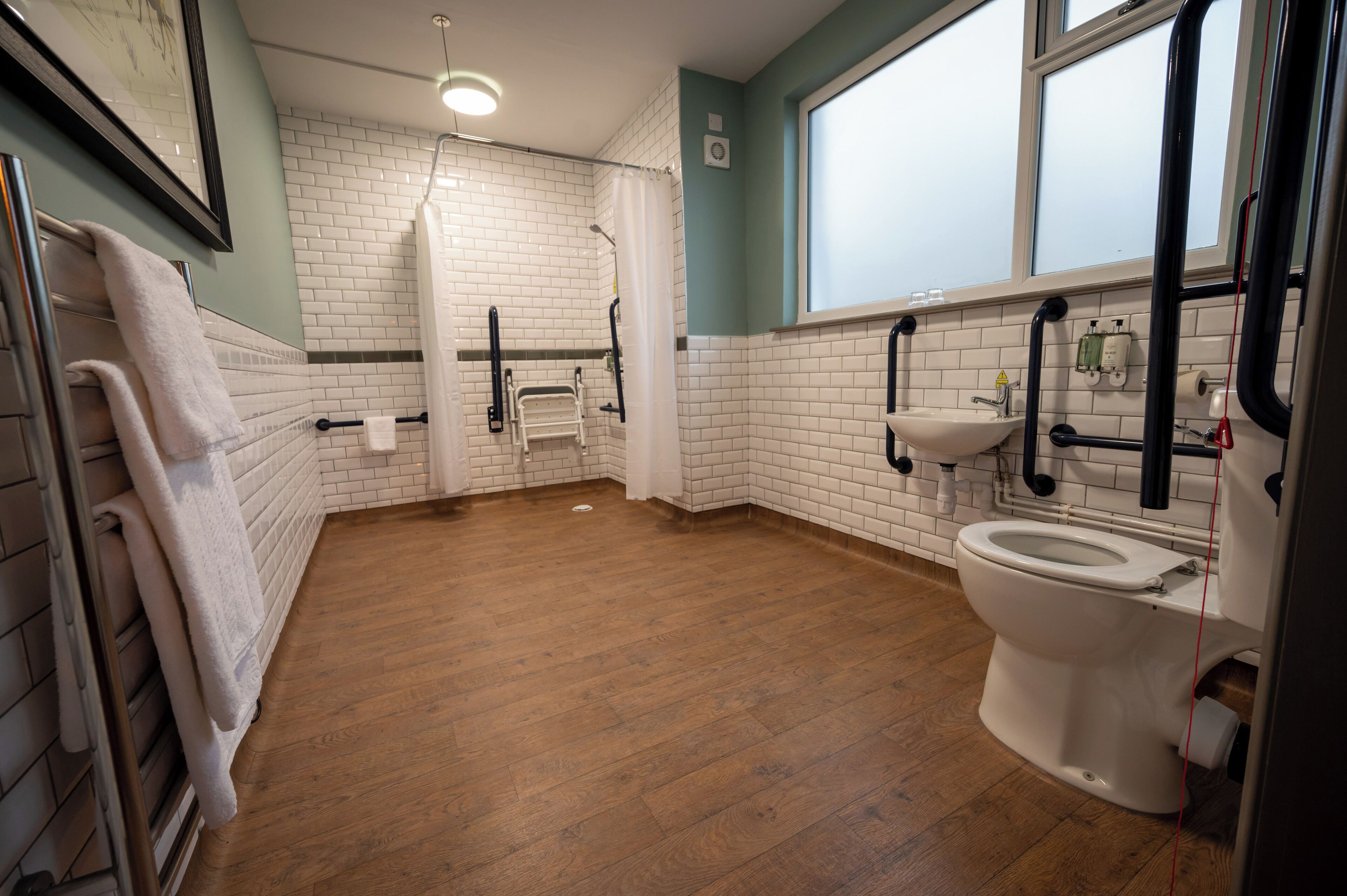 A bathroom with a wooden floor, white tiled walls, a white toilet, a sink, and a shower area.