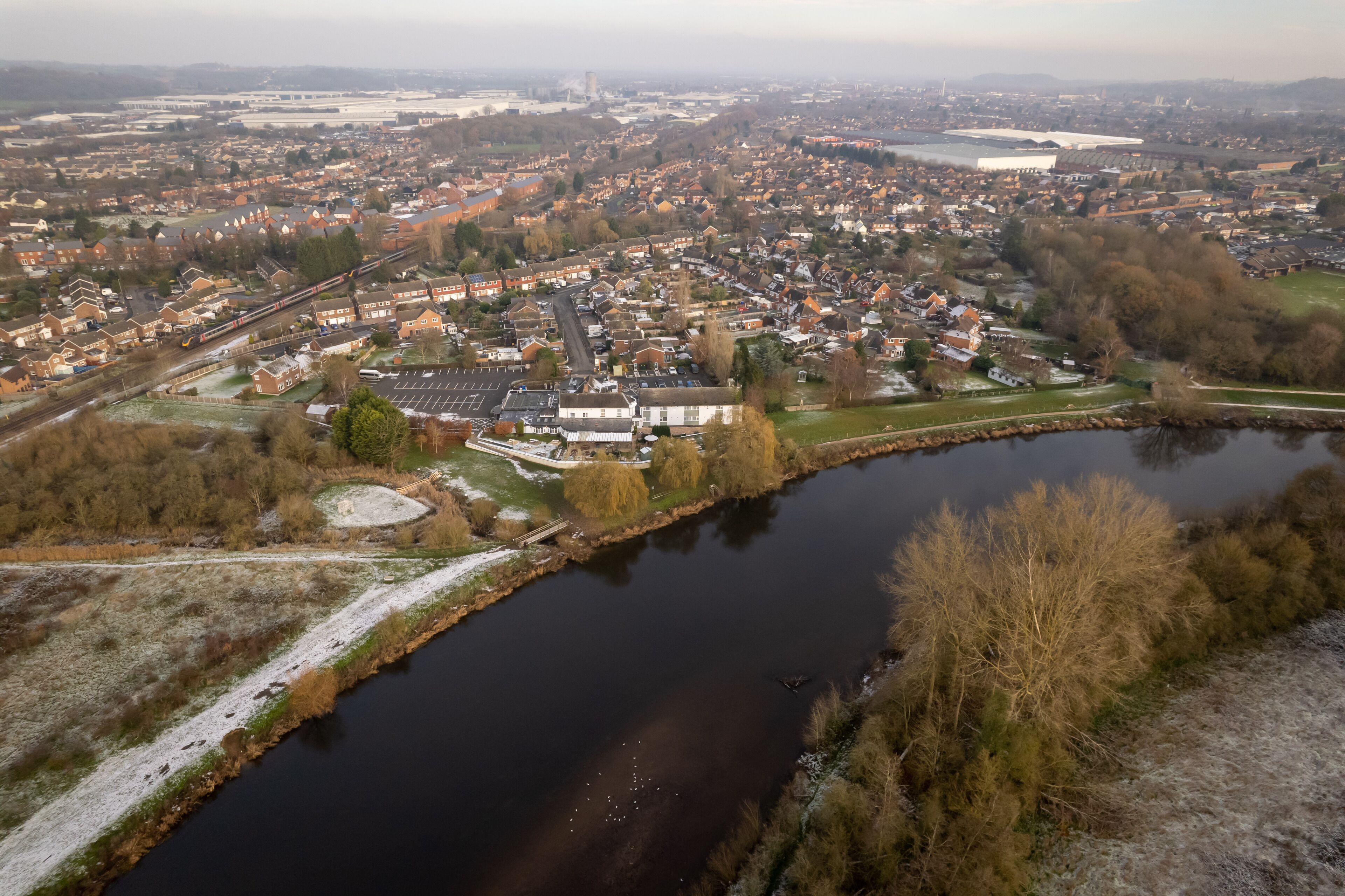 An aerial view of a town with a river, houses, and a large building with a prominent roof.