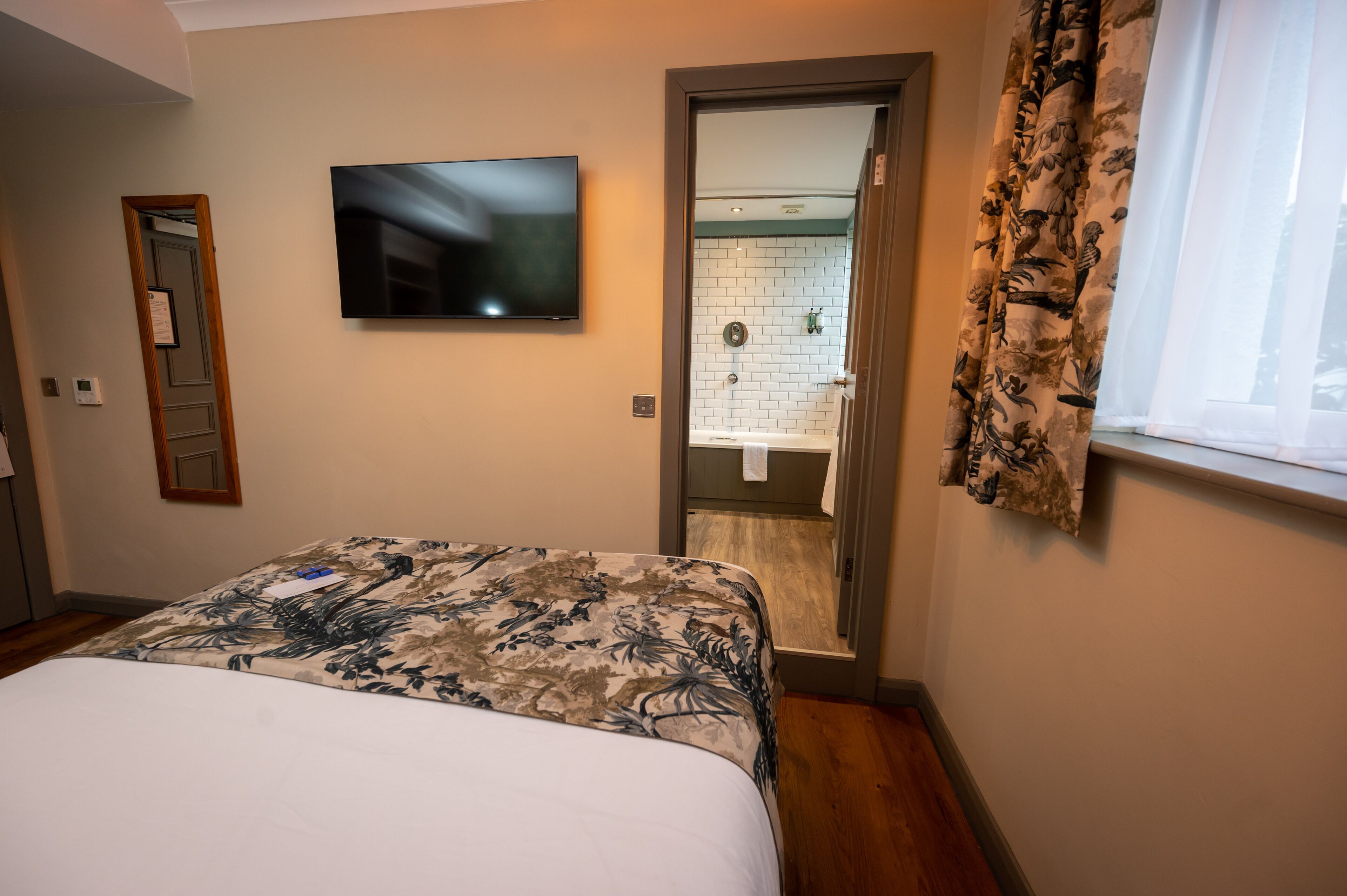 A hotel room with a bed, a television, a bathroom with a shower, and a window with curtains.