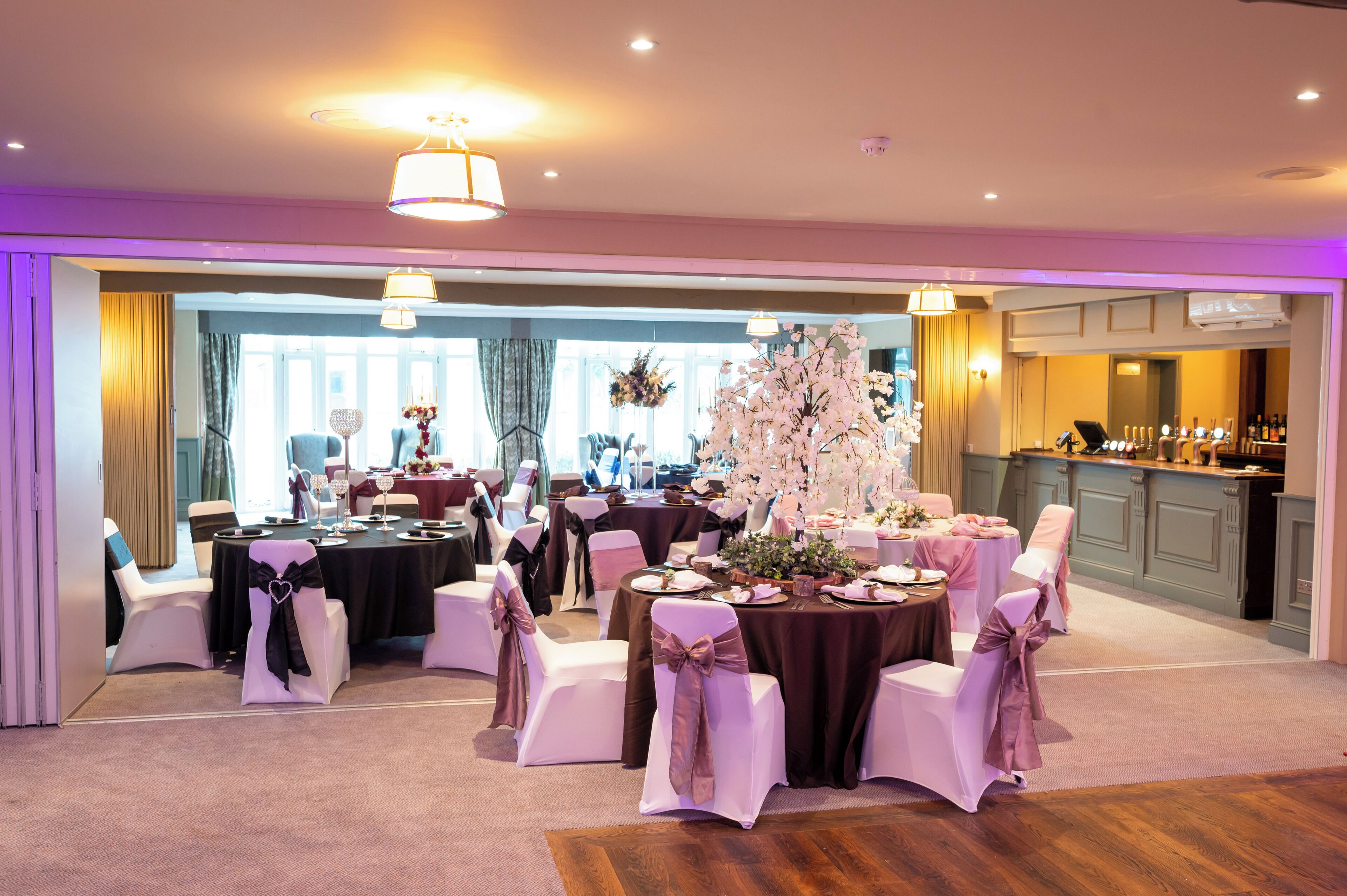 A banquet hall with round tables, white chairs, and pink sashes. There are chandeliers and a view of a kitchen area.