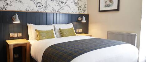 A neatly made bed with a plaid bedspread, two bedside lamps, and a floral wallpaper pattern.