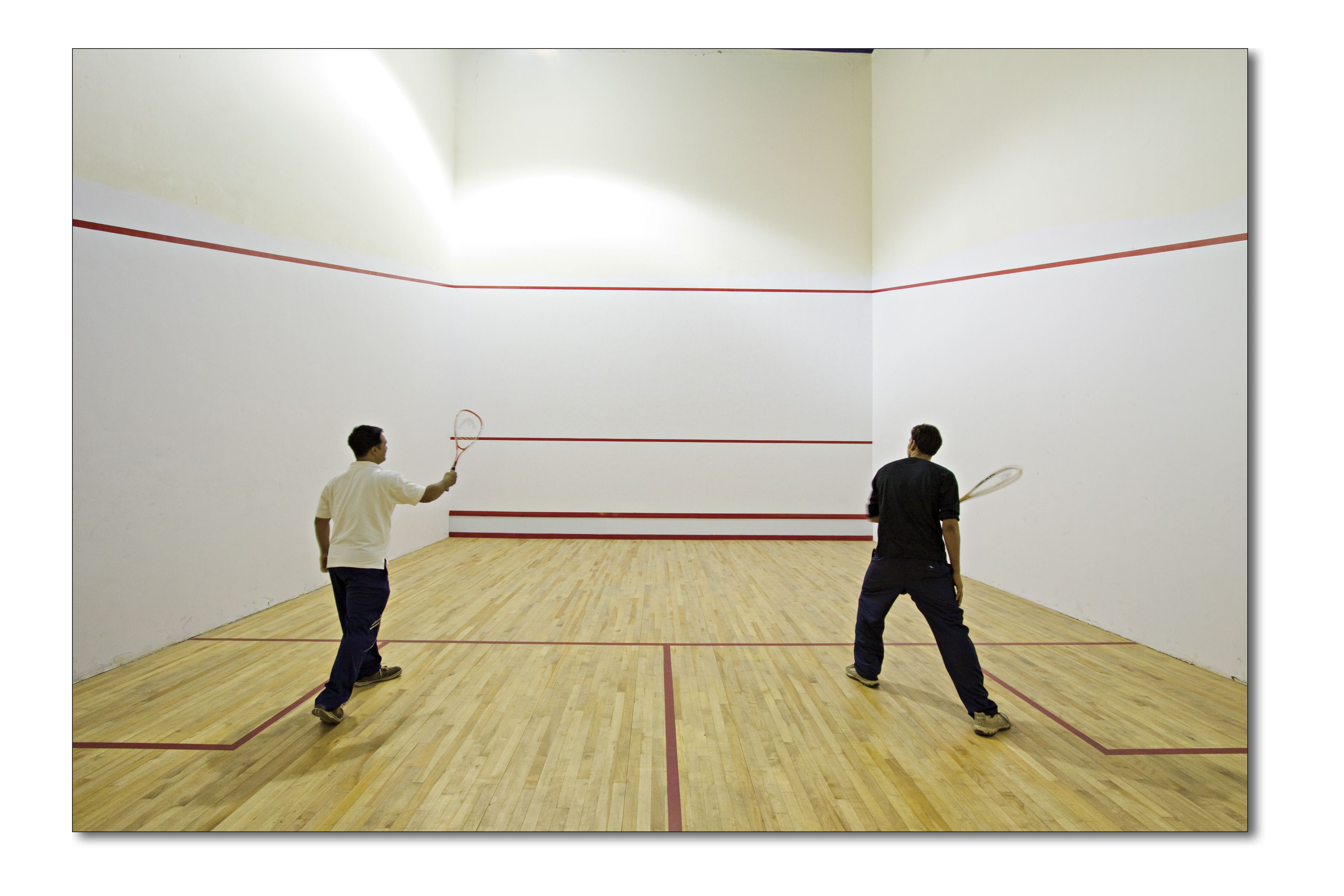 Two people playing squash in a room with wooden flooring and white walls.