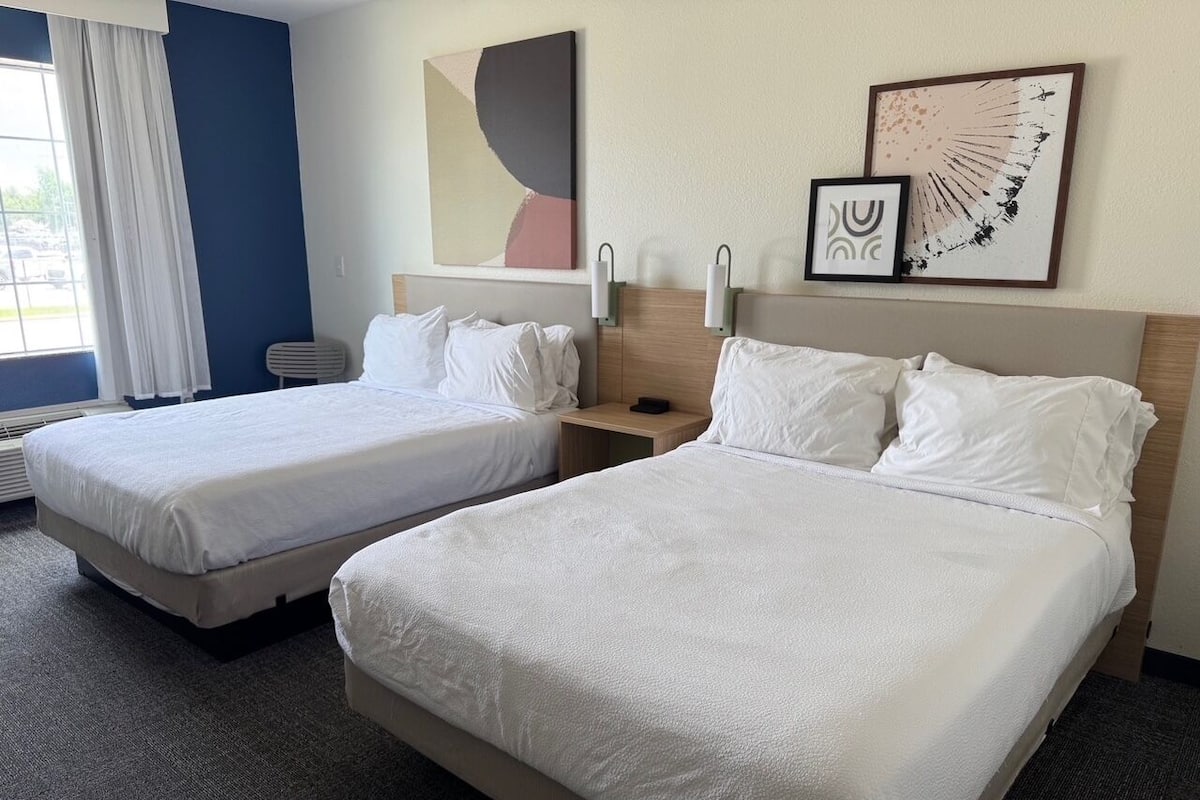 Two beds with white linens, a nightstand, wall art, and a window with curtains.