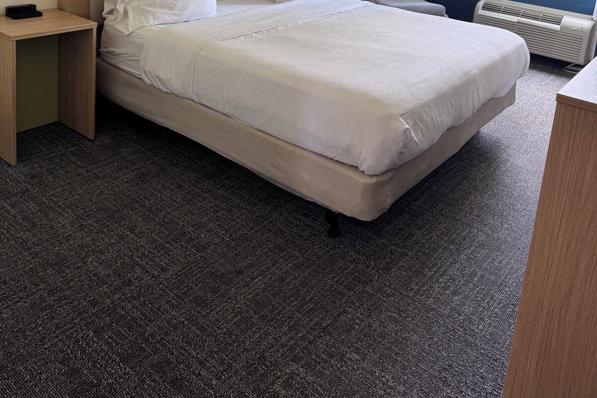 A hotel room with a large bed, a bedside table, a chair, a window with curtains, and a wall-mounted artwork.