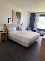 A hotel room with a large bed, a bedside table, a chair, a window with curtains, and a wall-mounted artwork.