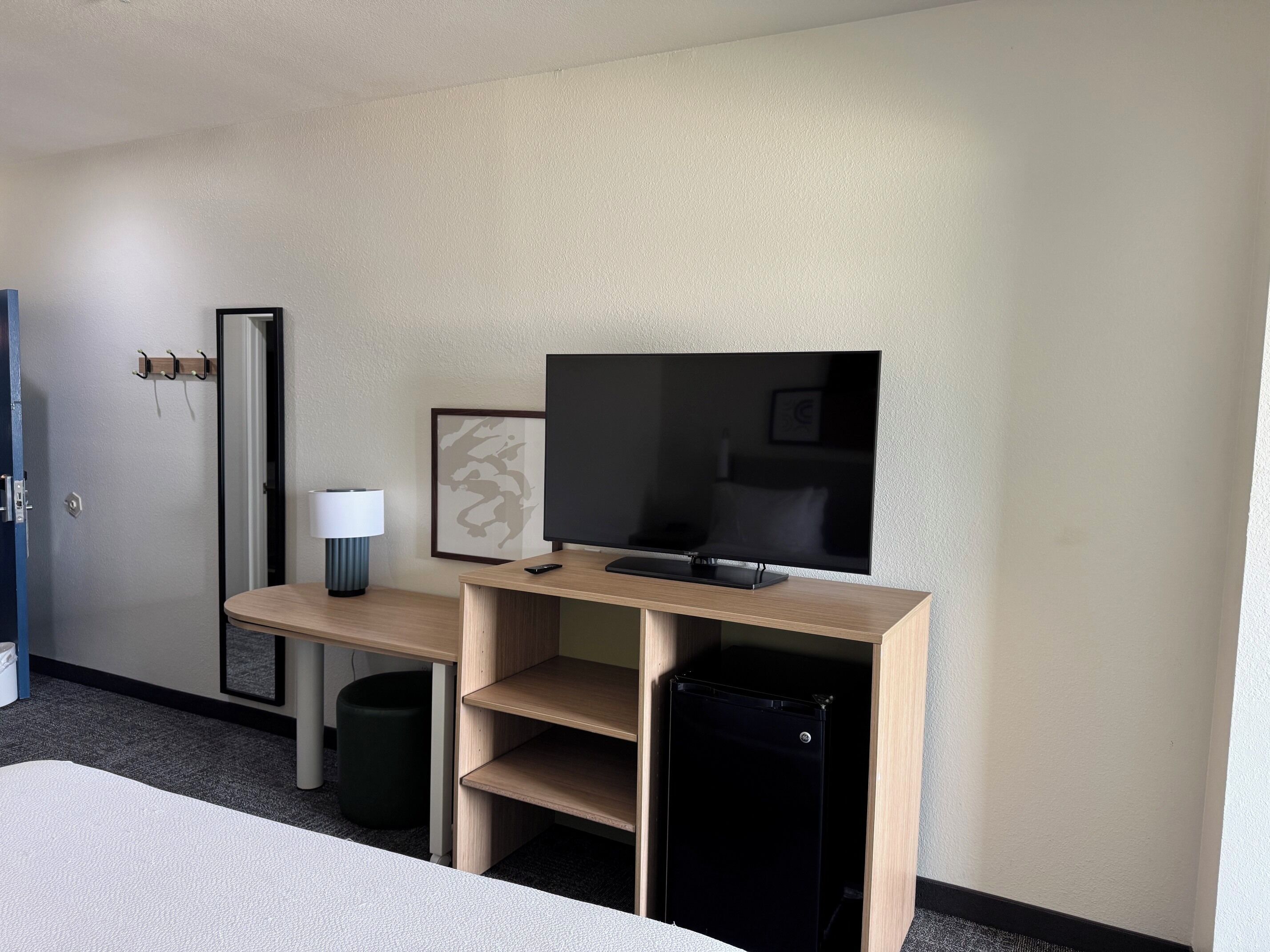 A hotel room with a bed, a desk with a television, a lamp, a framed picture, and a trash bin.