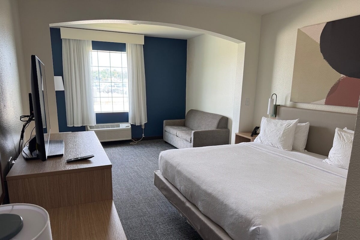 A hotel room with a bed, a desk, a chair, a TV, and a window with curtains.