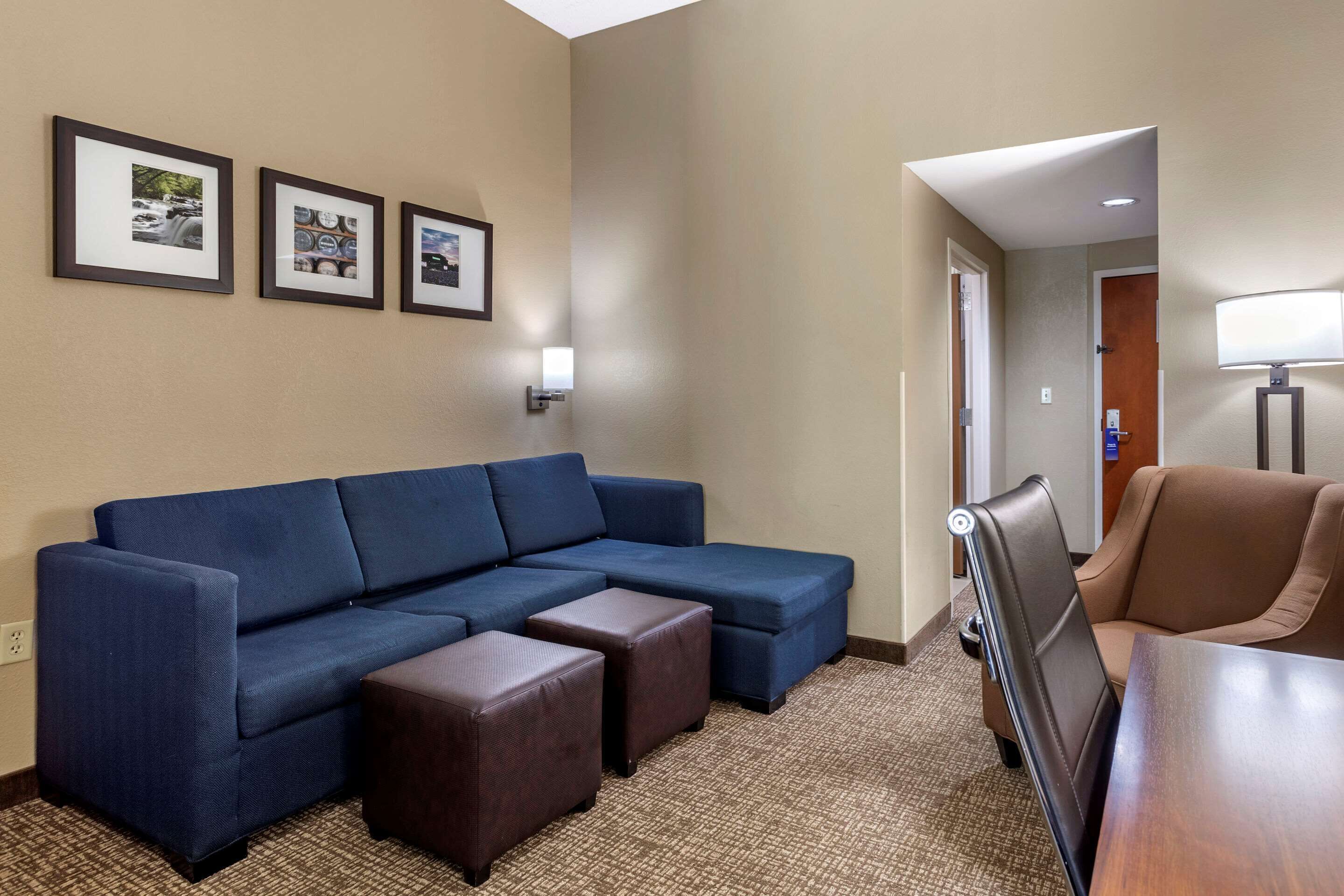 A hotel room with a sectional sofa, two ottomans, a brown armchair, a wooden table, and three framed pictures on the wall.