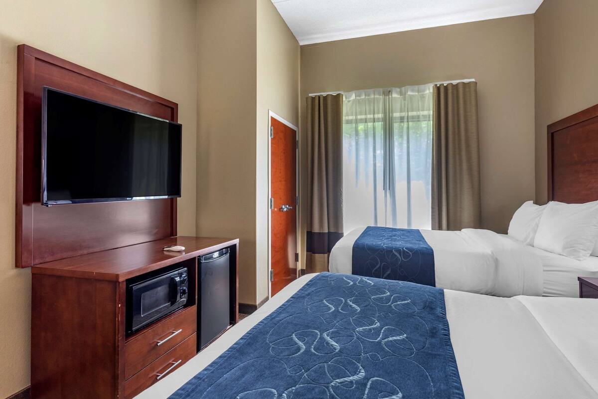 A hotel room with a TV, a desk with a microwave, two beds, and a window with curtains.
