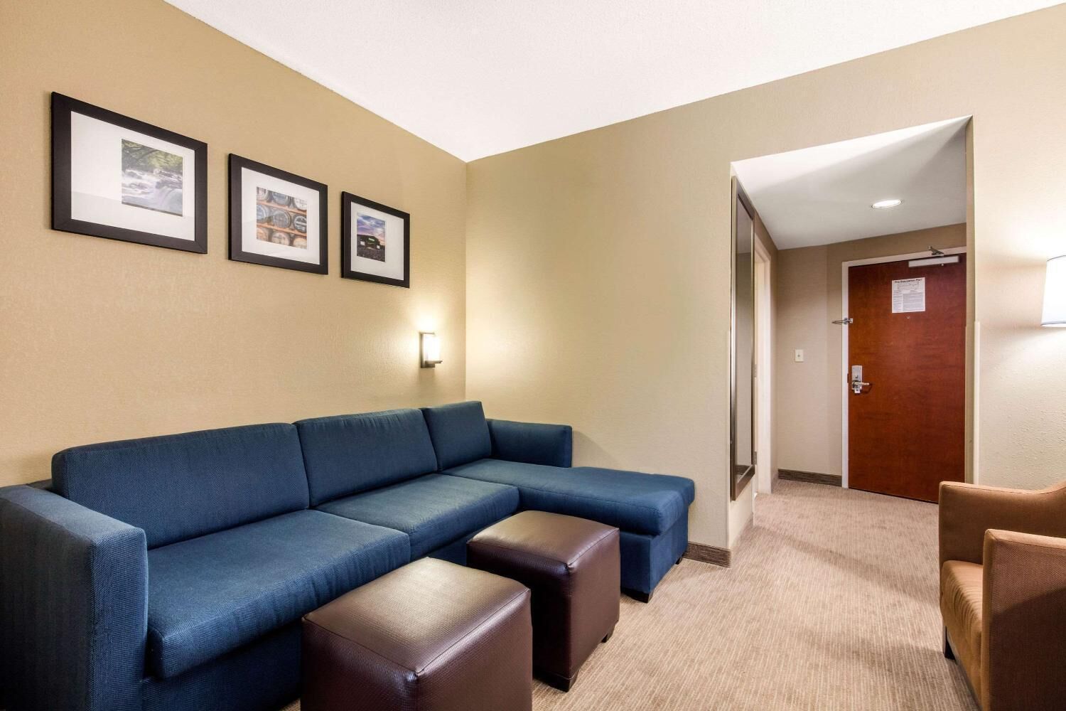 Habitación de hotel con un sofá seccional azul, un reposapiés marrón y un sillón marrón. Hay tres cuadros enmarcados en la pared y una puerta con un cartel.