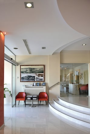 A modern hotel lobby with a curved staircase, a reception desk, and red chairs. - Blue Sea (Lesvos)