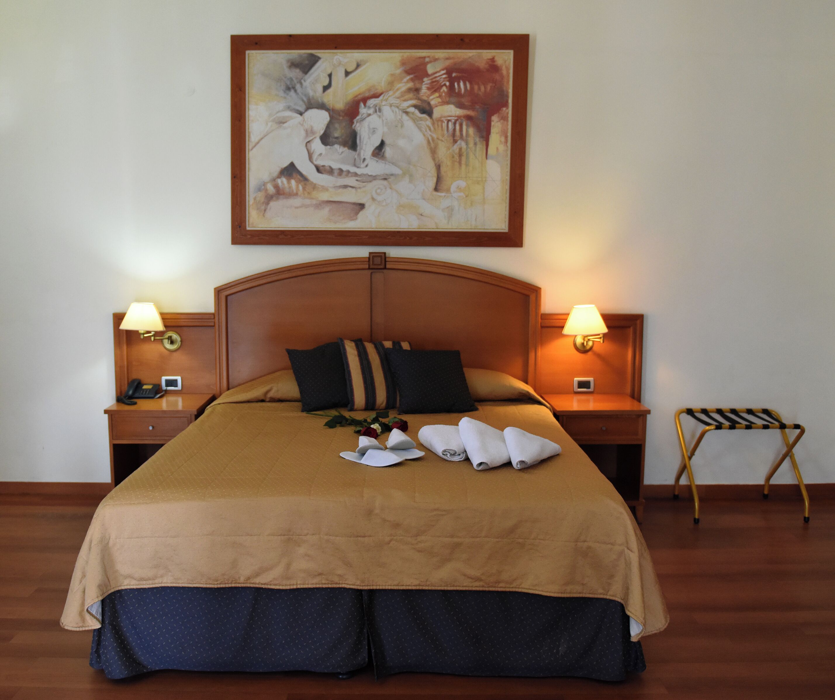 A hotel room with a large bed, two bedside tables with lamps, a wooden headboard, and a painting above the bed.