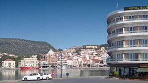 A multi-story hotel with balconies, a view of a historic town, and a body of water in the foreground. - Blue Sea (Lesvos)