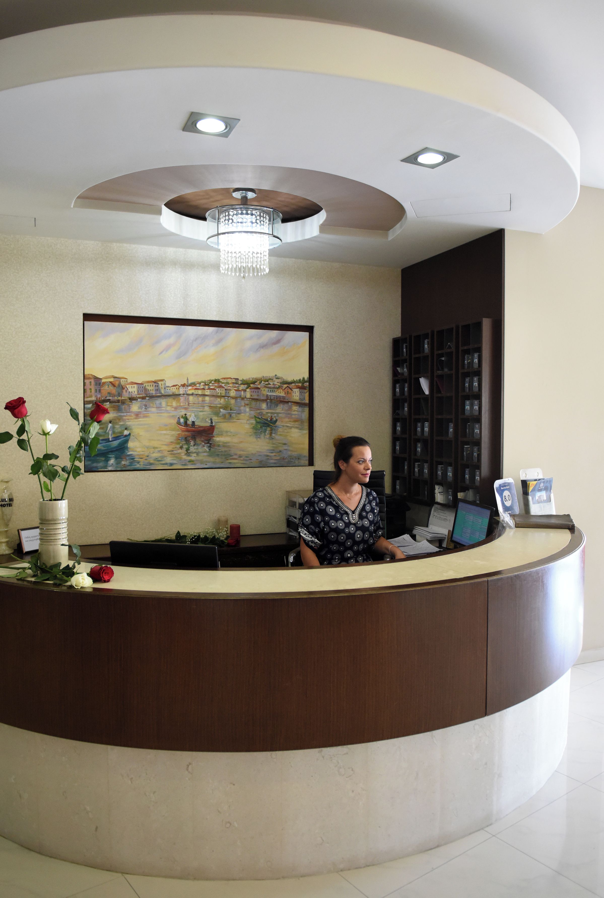 A reception desk with a painting of a harbor scene, a vase with flowers, and a chandelier.