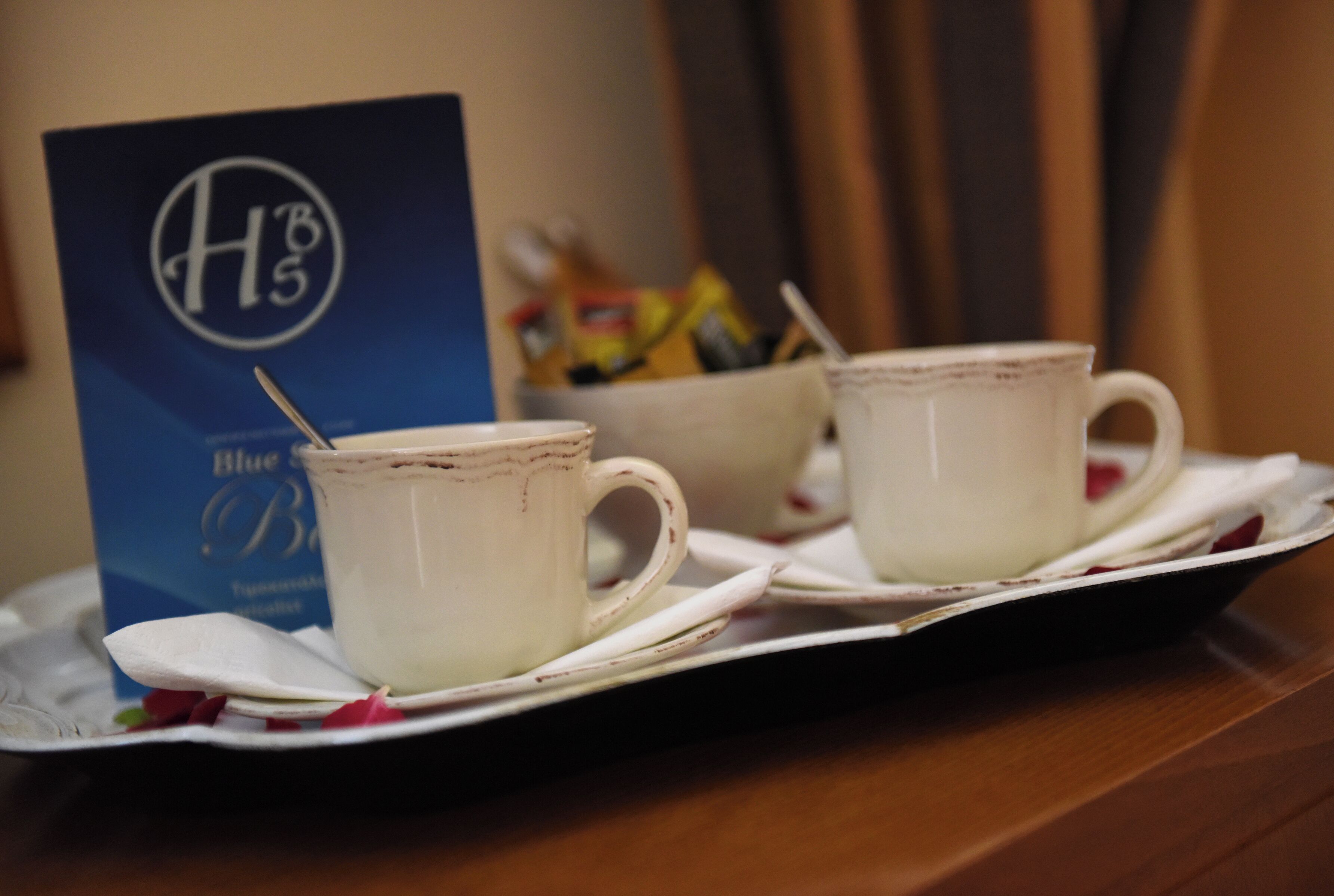 Two white ceramic cups on a tray with a spoon, a blue sign with 'HB5' logo, and a bowl of snacks in the background.