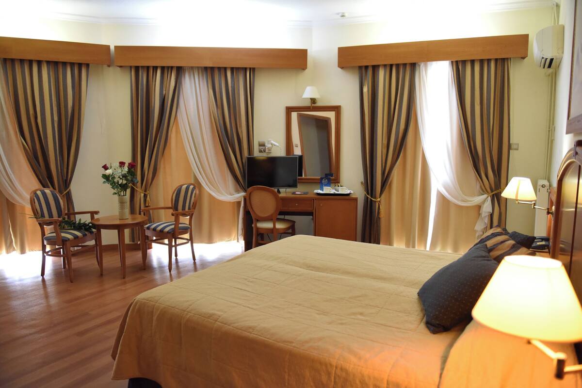 Une chambre d’hôtel avec un grand lit, un bureau avec un ordinateur, deux chaises, une petite table avec un vase de fleurs et des rideaux.