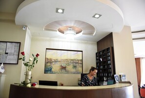 A reception desk with a vase of flowers, a painting of a canal scene, and a woman seated behind the desk. - Blue Sea (Lesvos)