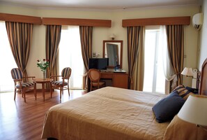 A hotel room with a large bed, a desk with a computer, two chairs, a small table with a vase of flowers, and curtains.