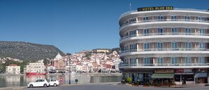 A multi-story hotel with balconies, a view of a historic town, and a body of water in the foreground. - Blue Sea (Lesvos)