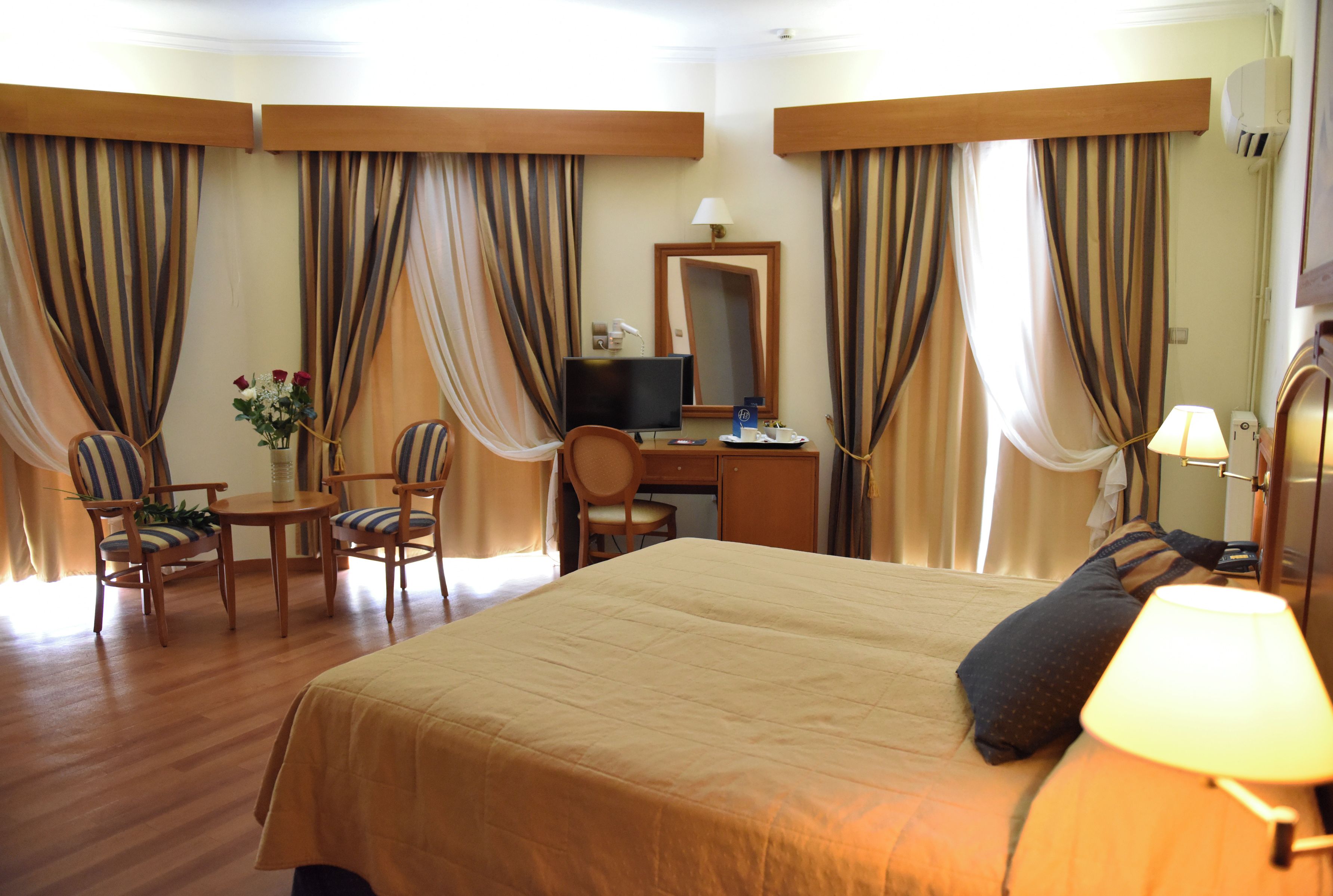 A hotel room with a large bed, a desk with a computer, two chairs, a small table with a vase of flowers, and curtains.