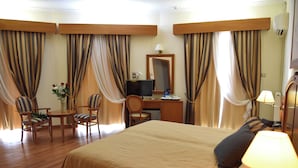 Une chambre d’hôtel avec un grand lit, un bureau avec un ordinateur, deux chaises, une petite table avec un vase de fleurs et des rideaux.