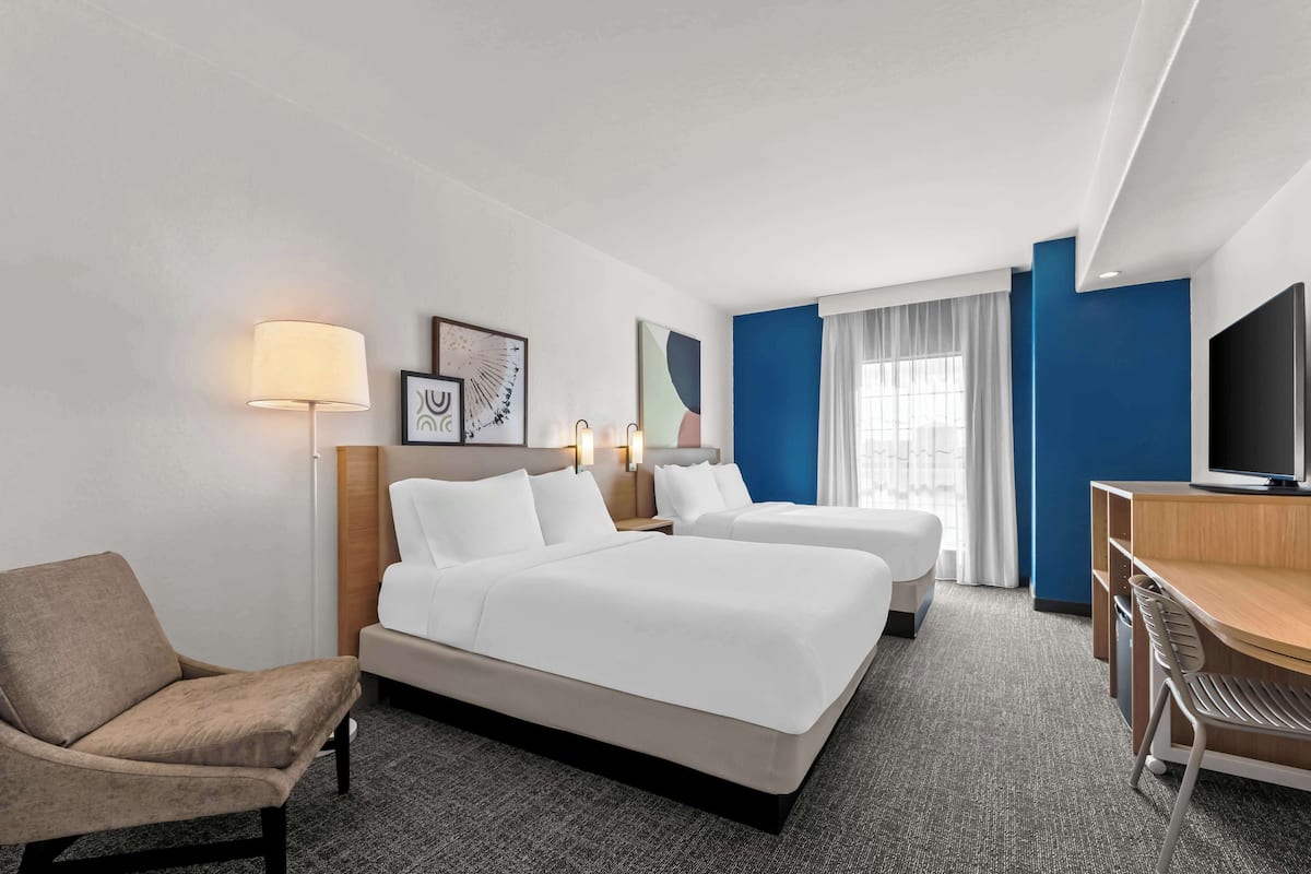 A hotel room with two single beds, a desk, a chair, a lamp, and a television.