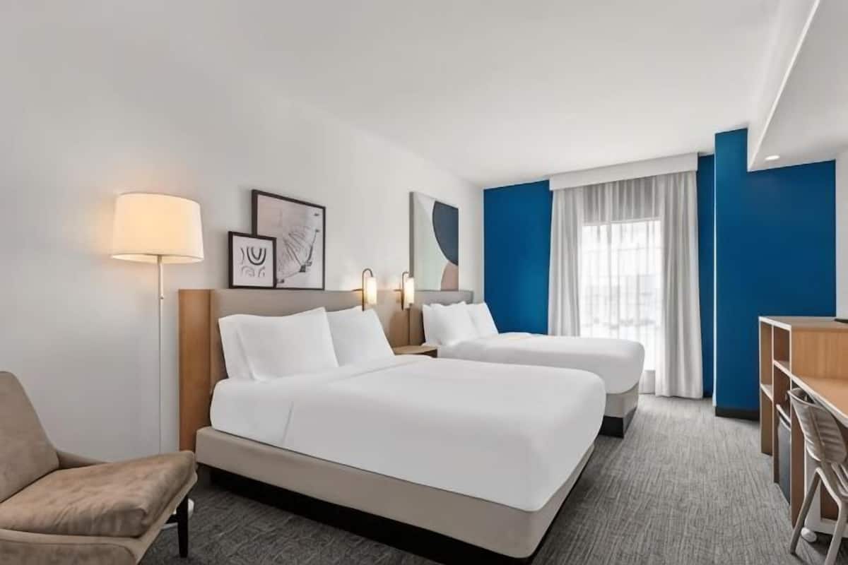 A hotel room with two beds, a desk, a chair, a lamp, and a window with curtains.