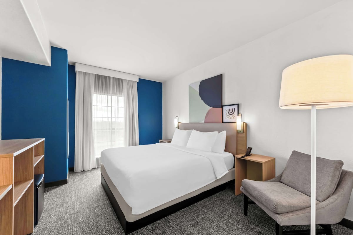 A hotel room with a bed, a desk, a chair, a lamp, and a painting on the wall.