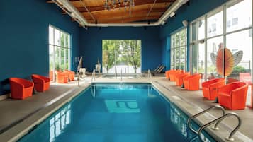 An indoor swimming pool with blue water, surrounded by orange chairs, large windows, and a wooden ceiling.