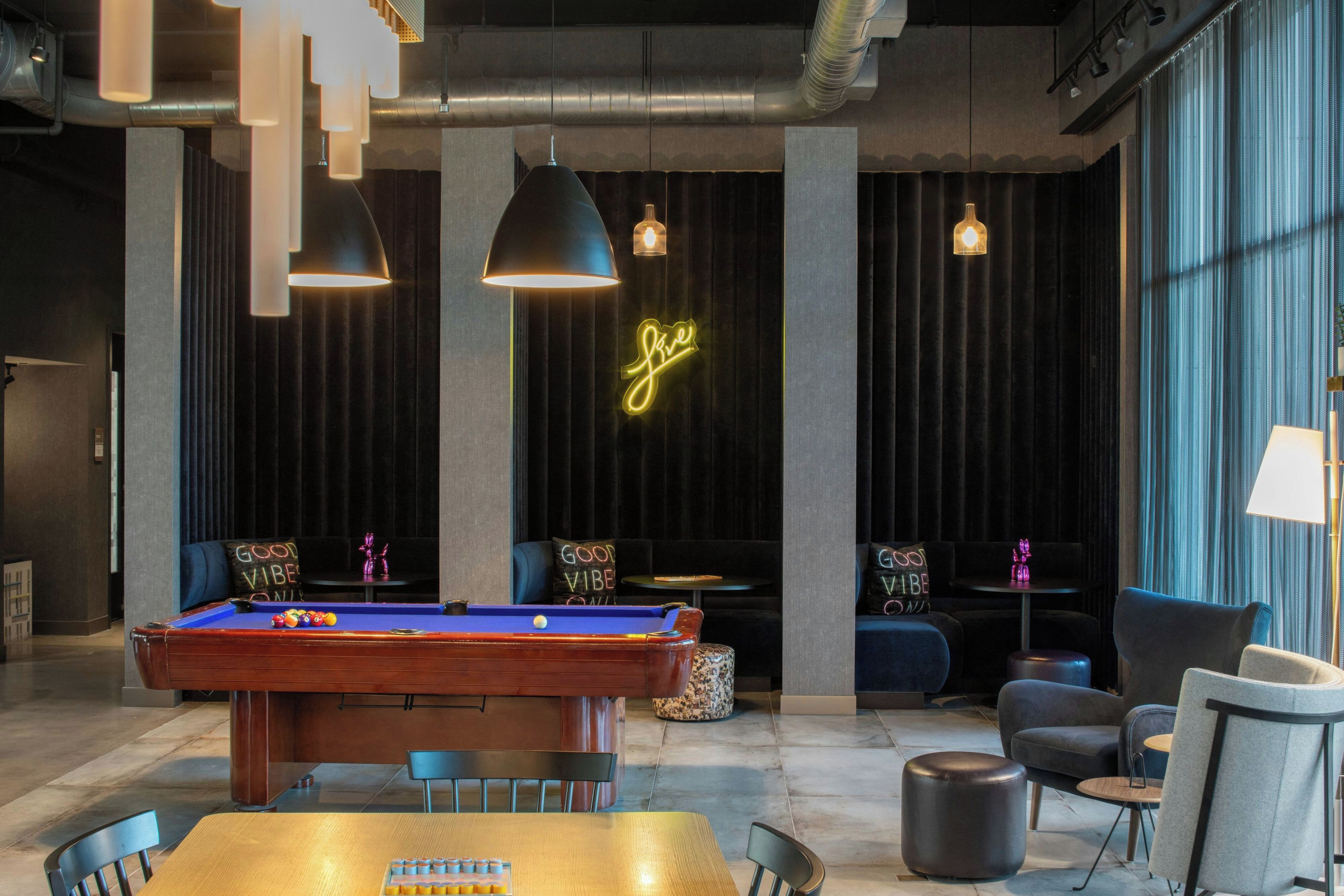 A billiards table with a pool cue, chairs, and a neon sign in a modern lounge area with dark curtains and pendant lighting.