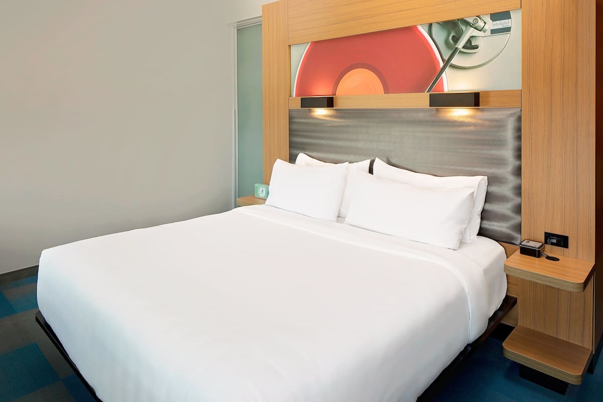 A neatly made bed with white linens, a wooden headboard with a built-in light, and a small bedside table with a clock.