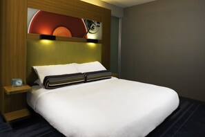 A neatly made bed with white linens and striped pillows, a bedside table with a clock, and a wall-mounted light fixture. - Aloft by Marriott Green Bay (Green Bay)