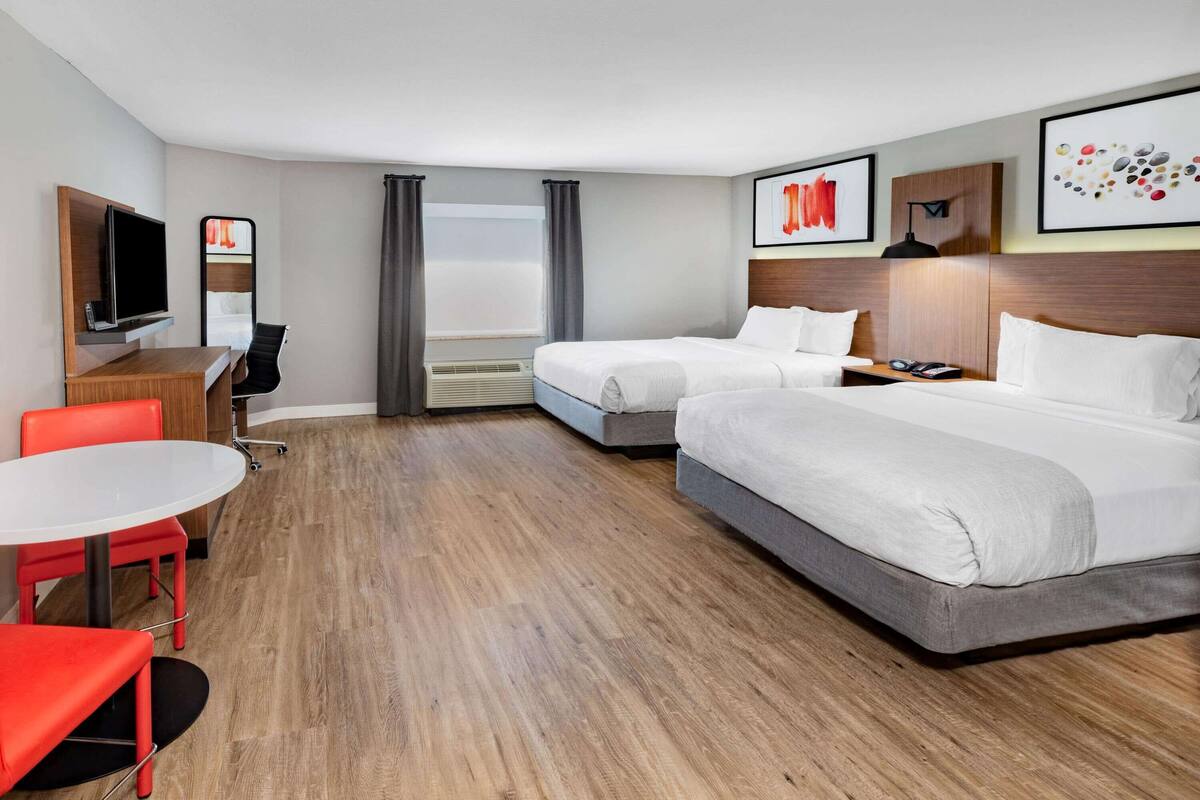 A hotel room with two beds, a desk with a TV, a small table, and red chairs.