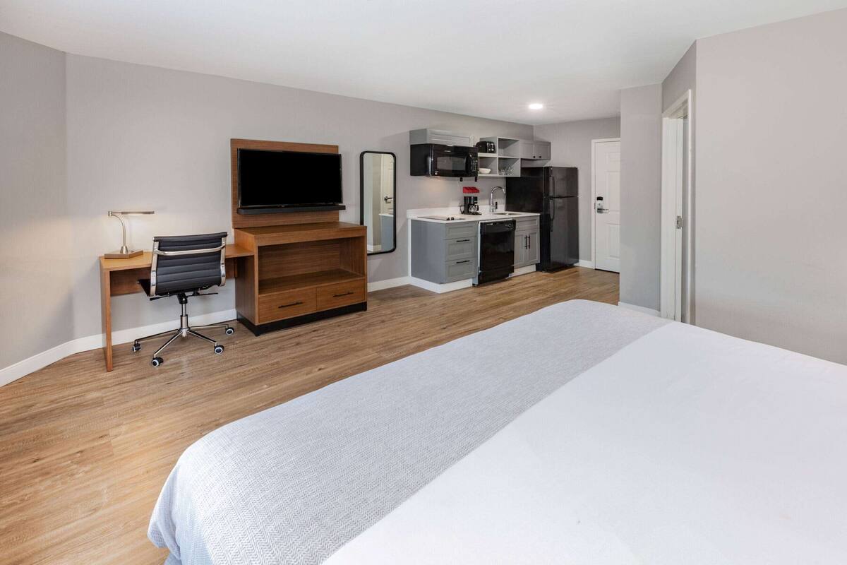 A modern hotel room with a large bed, a desk with a chair, a flat-screen TV, a kitchenette with a microwave and refrigerator, and a bathroom door.