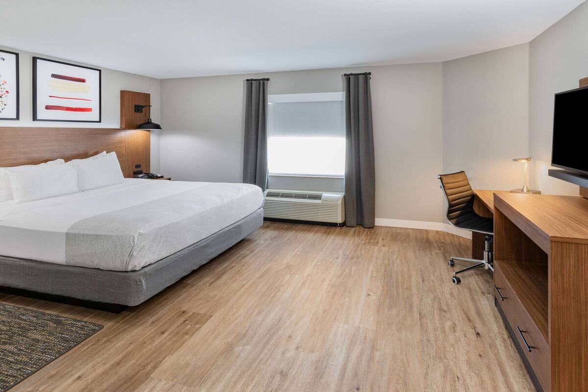 A hotel room with a bed, desk, chair, TV, air conditioning unit, and curtains.