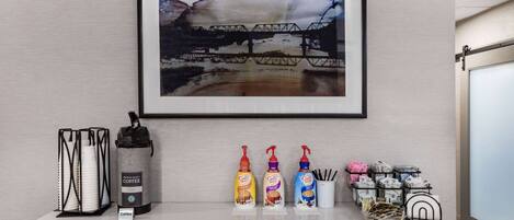 A hotel reception desk with a framed artwork of a bridge, a coffee station with branded coffee dispensers, and neatly arranged mail and magazines.