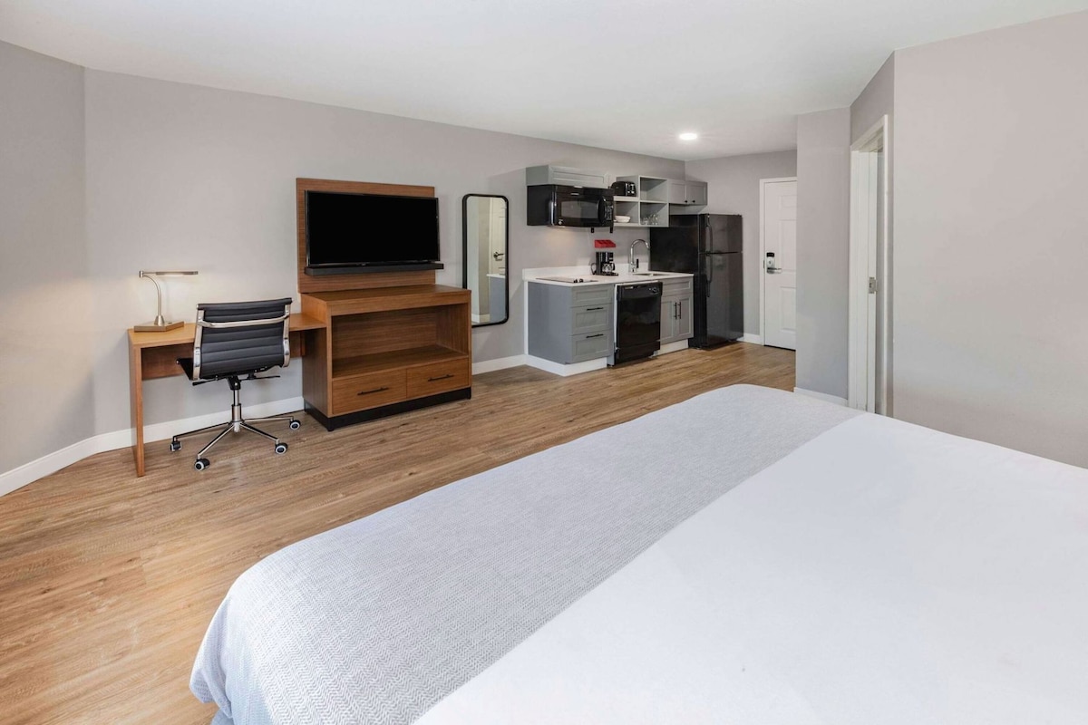 A hotel room with a bed, a desk with a chair, a TV, a kitchenette, and a mirror.