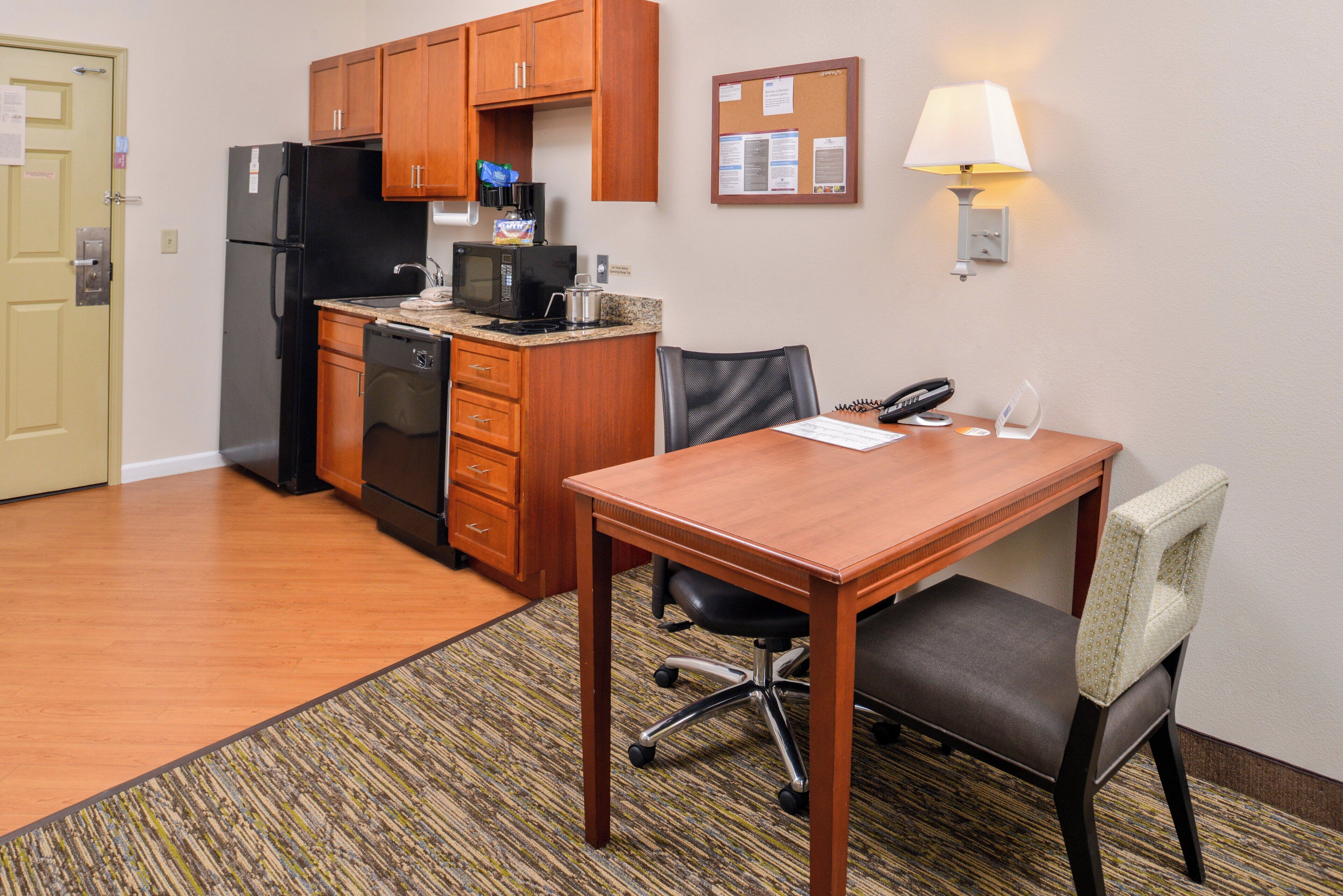 A compact hotel room with a kitchenette, a dining table with a chair, a desk with a chair, a lamp, and a bulletin board.
