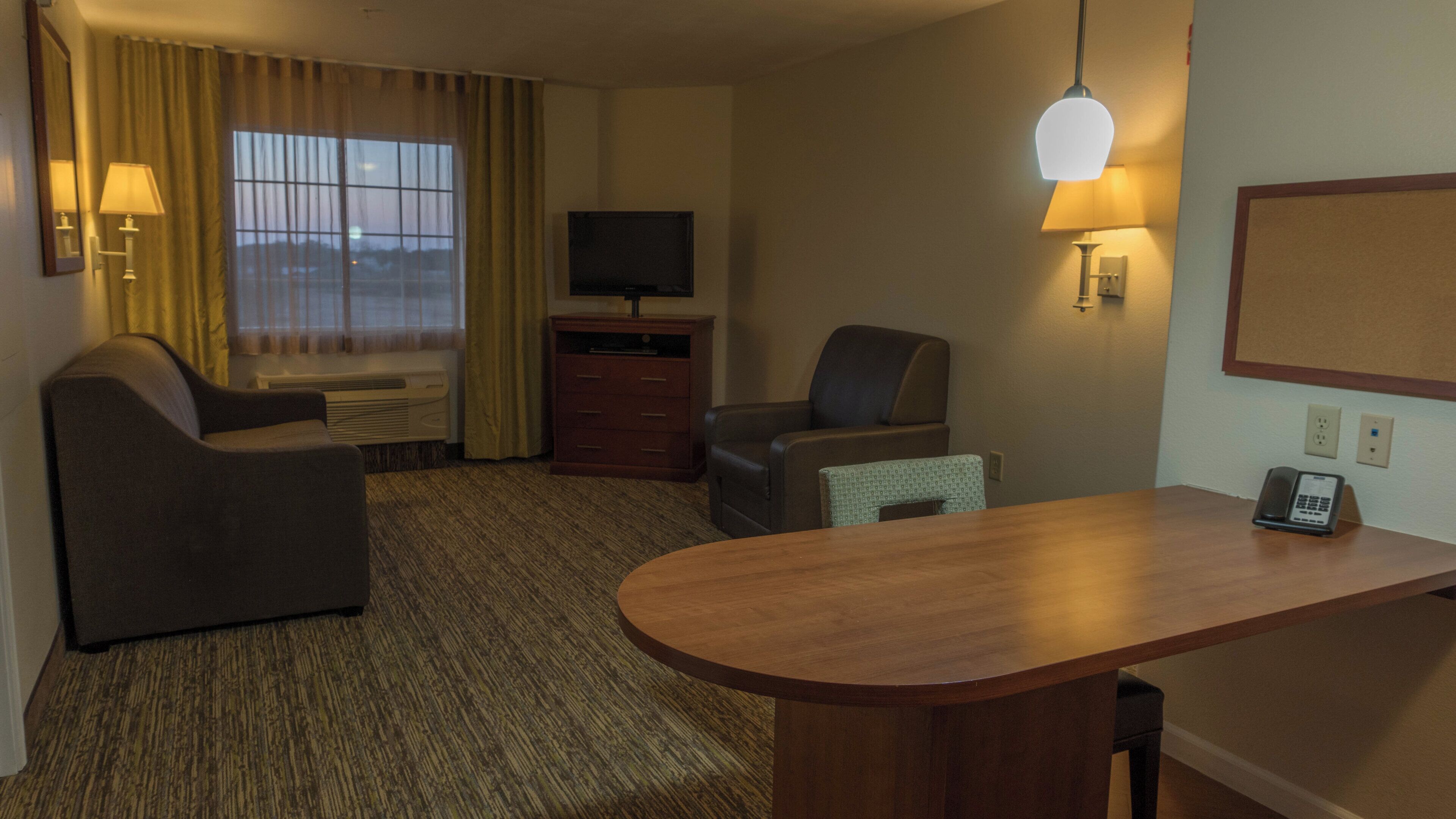 Ein Hotelzimmer mit Schreibtisch, zwei Stühlen, einem Fernseher, einer Lampe und einem Fenster mit Vorhängen.