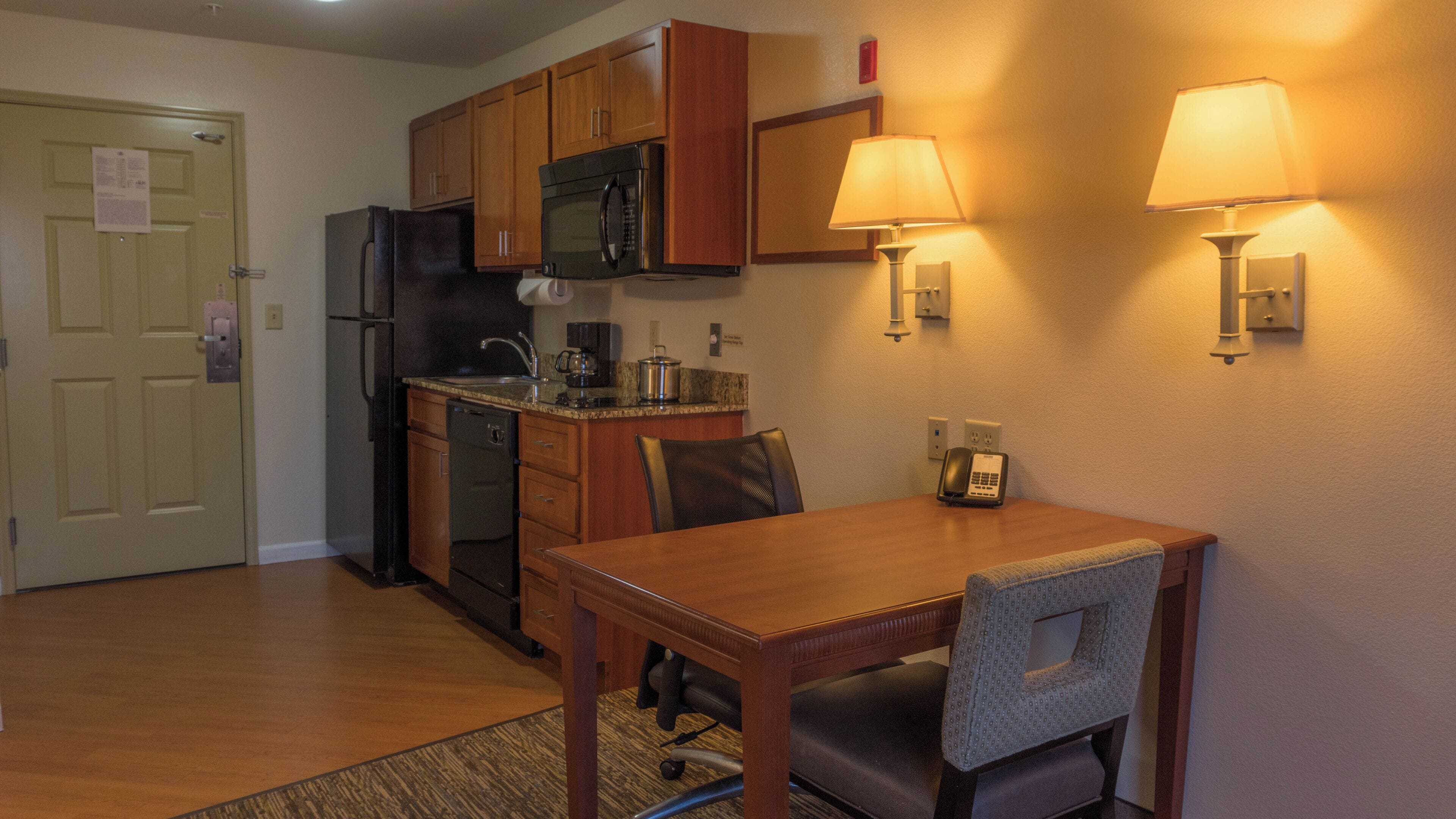 A compact kitchen with wooden cabinets, a microwave, and a refrigerator. There is a dining table with chairs and two wall-mounted lamps.