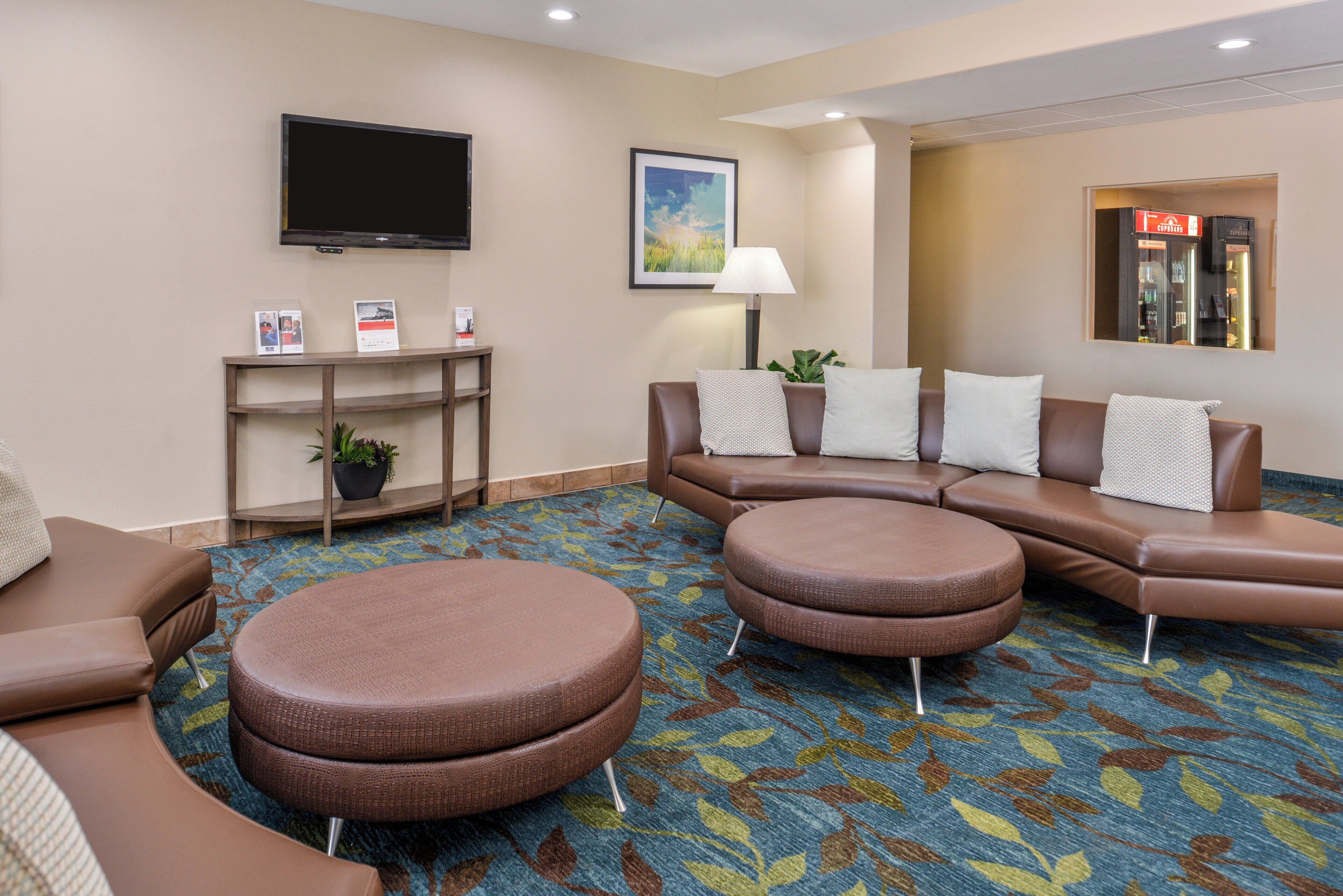 A hotel lobby with a brown leather sofa, two round ottomans, a flat-screen TV, a framed landscape painting, a lamp, and a small table with brochures.