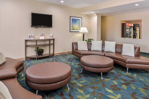 A hotel lobby with a brown leather sofa, two round ottomans, a flat-screen TV, a framed landscape painting, a lamp, and a small table with brochures. - Candlewood Suites ABILENE by IHG (Abilene)
