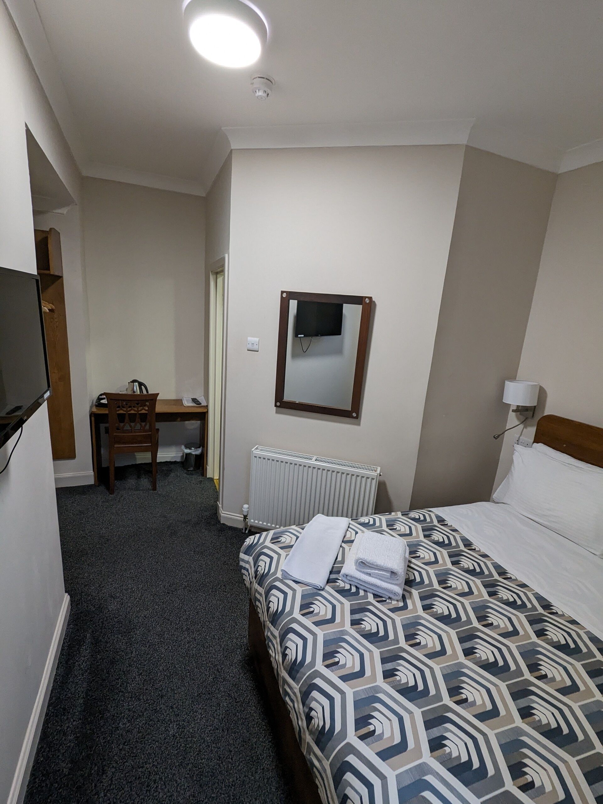 A hotel room with a bed, a desk, a chair, a television mounted on the wall, and a radiator.