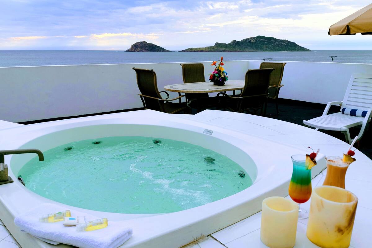 A hot tub with a view of the sea, a table with chairs, and two drinks with fruit garnishes.